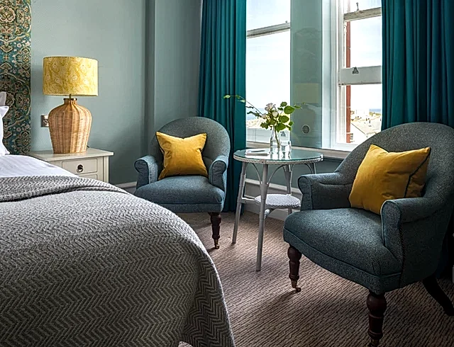 Luxury coastal hotel room in Newquay with turquoise armchairs, mustard-yellow cushions and a large patterned headboard. The sunlit suite features a glass bistro table and views of the Cornish coast.
