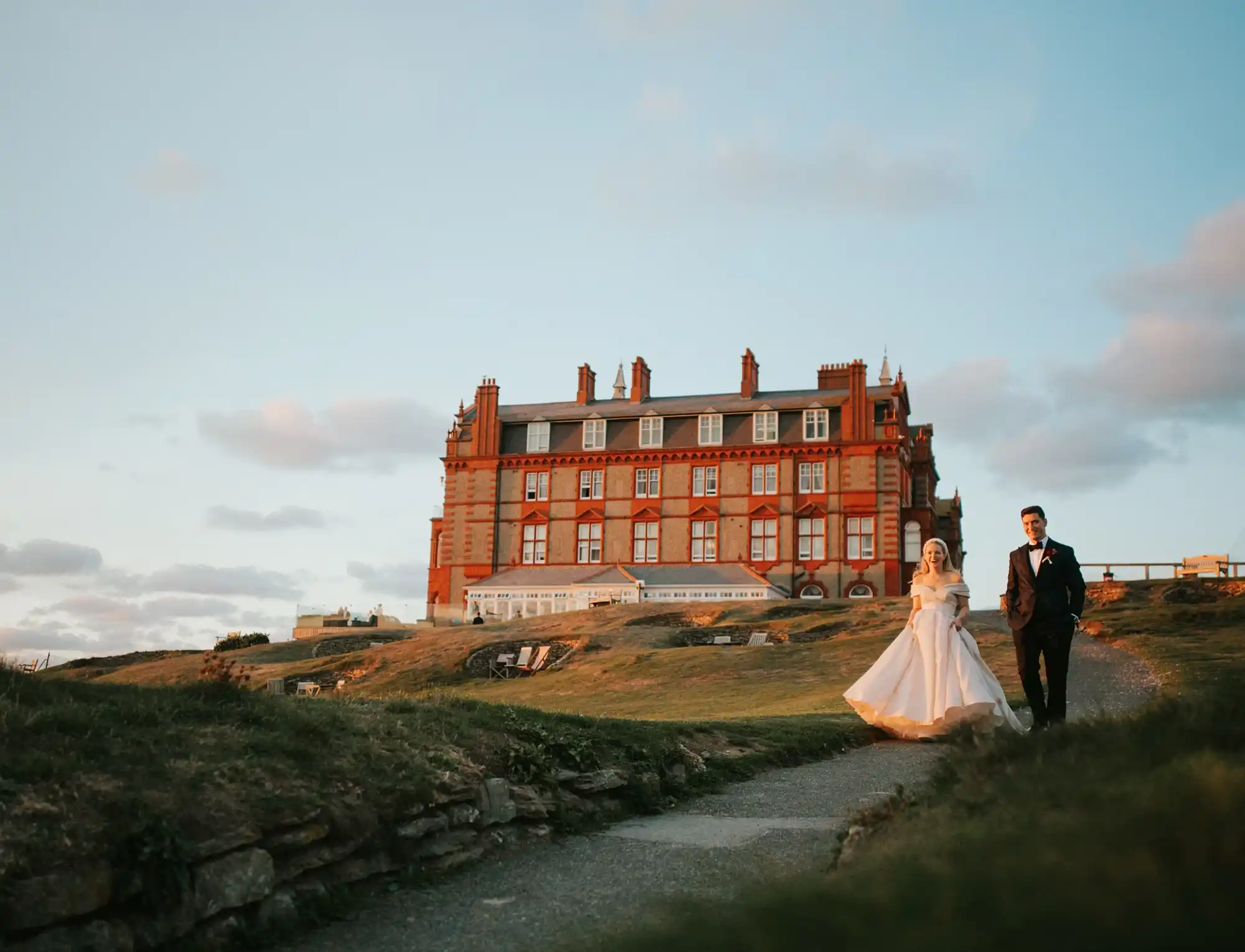 Bride and groom walking down the grass cliffs outside The Headland Hotel in Newquay. The bride is wearing a long white wedding dress and veil and the groom is wearing a black and white suit with a black bow tie. 