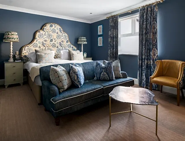 Luxury boutique-style hotel room in Newquay featuring deep blue walls and a statement scalloped headboard in floral fabric. This coastal suite includes a navy and yellow accent sofa, a marble coffee table and a mustard-yellow armchair by a window with traditional patterned drapes.
