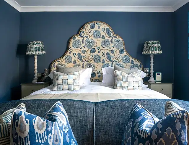 A modern, cosy hotel room painted in a deep blue colour, including a traditional, large, scalloped headboard with floral fabric. Identical lamps sit on either side of the bed on white, wooden bedside tables. patterened duck-feather pillows are placed on the bed and sofa. 