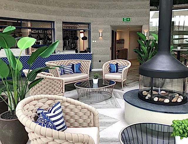 The Deck Restaurant within The Headland Hotel, features an open kitchen and bar with blue tiles and beige walls. In the centre of the room, there is a black circular fireplace. 