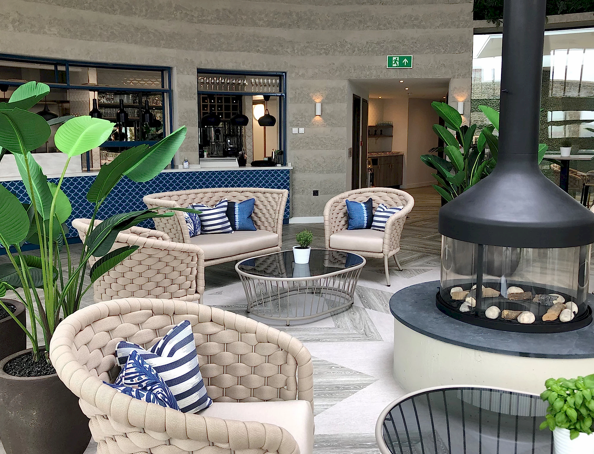 The Deck Restaurant within The Headland Hotel, features an open kitchen and bar with blue tiles and beige walls. In the centre of the room, there is a black circular fireplace.