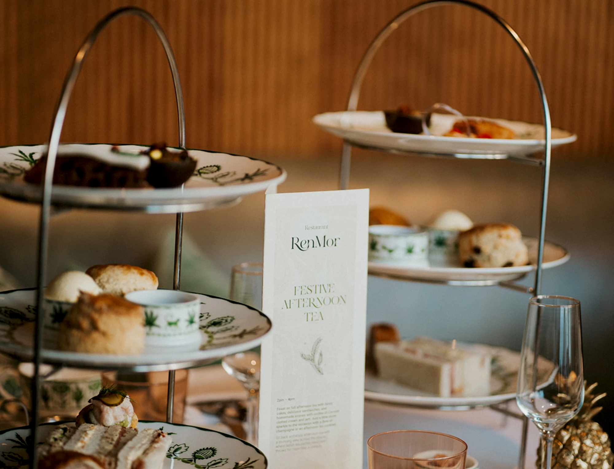 Two RenMor Afternoon Tea stands featuring various cakes, sandwiches and scones with a pot of jam and cream. 