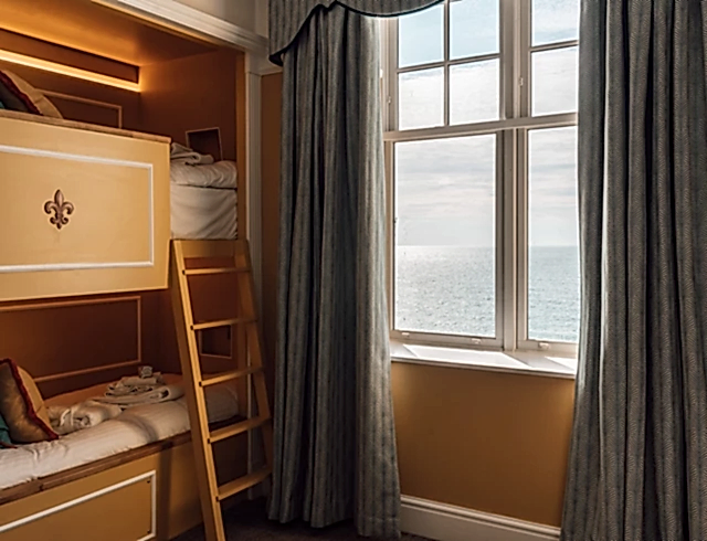 Luxury children’s hotel suite with warm orange walls and a custom wooden bunk bed. A large window offers stunning Atlantic Ocean views, framed by elegant teal patterned curtains in a traditional style.