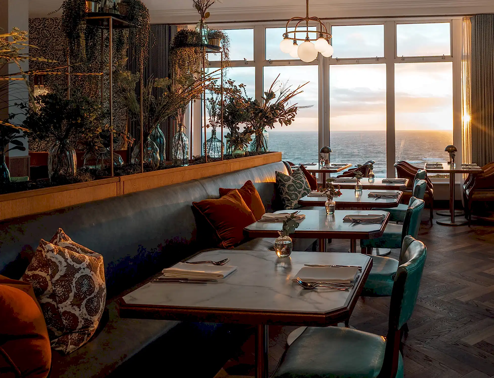 Luxury sea view restaurant at sunset with marble tables and botanical decor. Teal and wooden seats. 