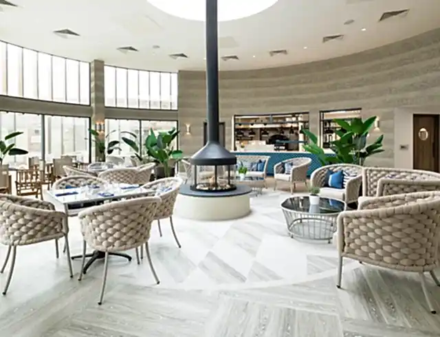 A restaurant featuring a modern circular fireplace, woven designer seating and bright floor-to-ceiling windows with coastal views.