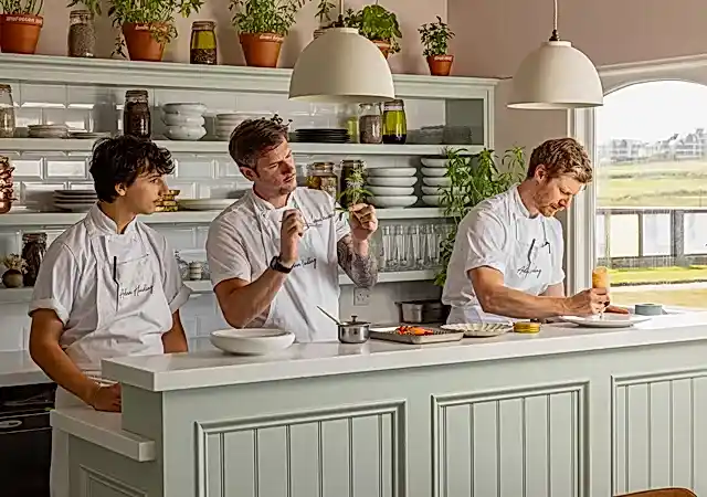 Three chefs are preparing food in an open kitchen surrounded by potted green plants and shelves stacked with crockery. A window within the kitchen area overlooks a golf course and various buildings. 