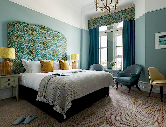 Luxury oceanfront hotel bedroom featuring a bespoke teal and yellow patterned headboard, plush designer soft furnishings and a floor-to-ceiling window with coastal views in Newquay.