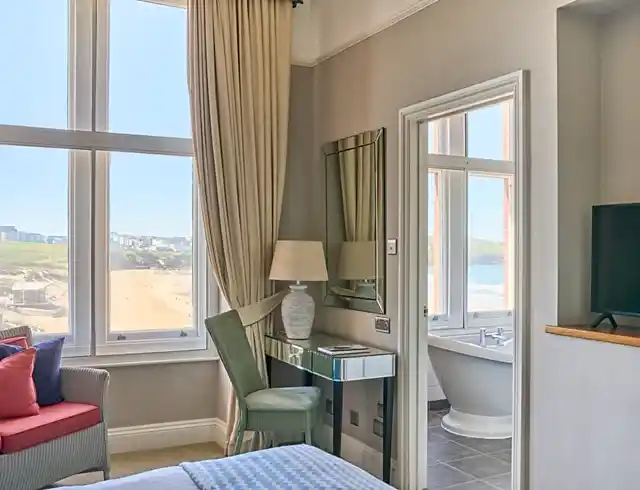 A bright hotel suite with a large window overlooking a beach, featuring a mirrored vanity, a green chair and a view into a bathroom with a freestanding tub.