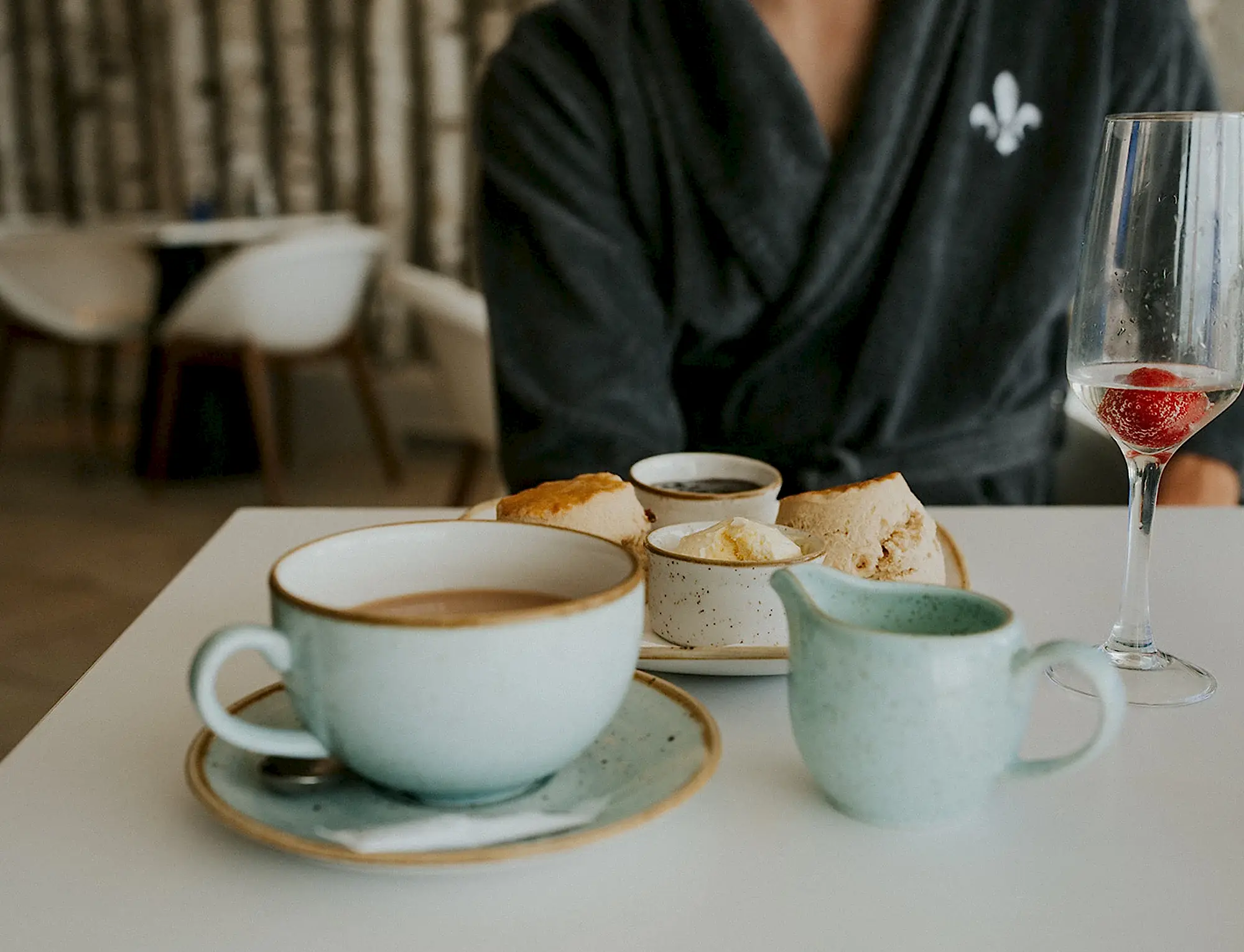 Cream Tea alongside a blue mug of tea and a glass of prosecco. Spa guest is dining in a grey Headland Spa robe. 