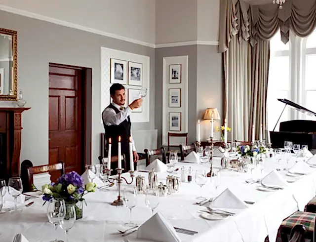 A waiter standing over a full length dining table with a grand piano in the background