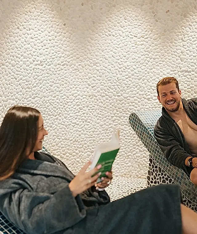 Two spa day guests sat on blue and white tiled tepidarium beds. One person is holding an open book and they are both looking and smiling at each other. Both people are wearing grey robes. 