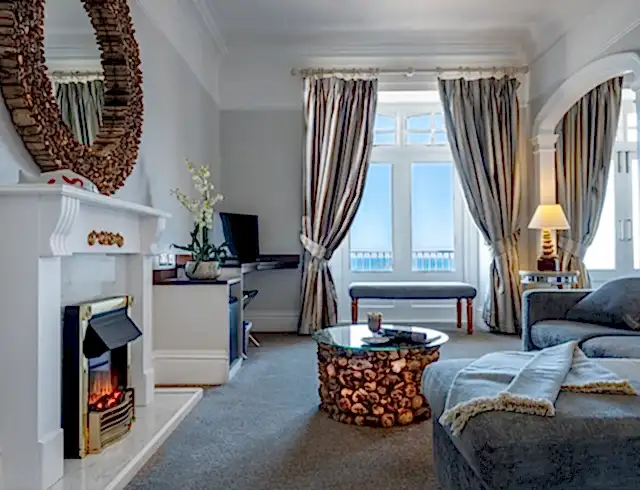 Luxury hotel bedroom at The Headland Hotel in Newquay with a circular wooden mirror above a fireplace, cosy grey sofa and open curtains showing a view of blue sky and the sea. 