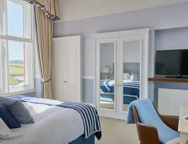 Luxury bed opposite mirrored wardrobe doors. A TV sits above a floating wooden shelf on a lilac-painted wall. Views out of the window show a lush green golf course and Cornish coastline.  