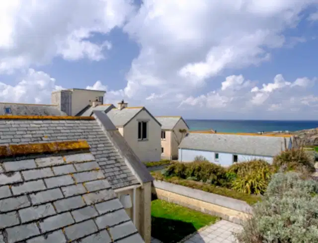 Image taken from a coastal multi-story cottage window showing views of the other blue and yellow cottages. Views of the Cornish coast and ocean are on the horizon. Green gardens can be seen outside cottages along with bushes and plants around the area. 