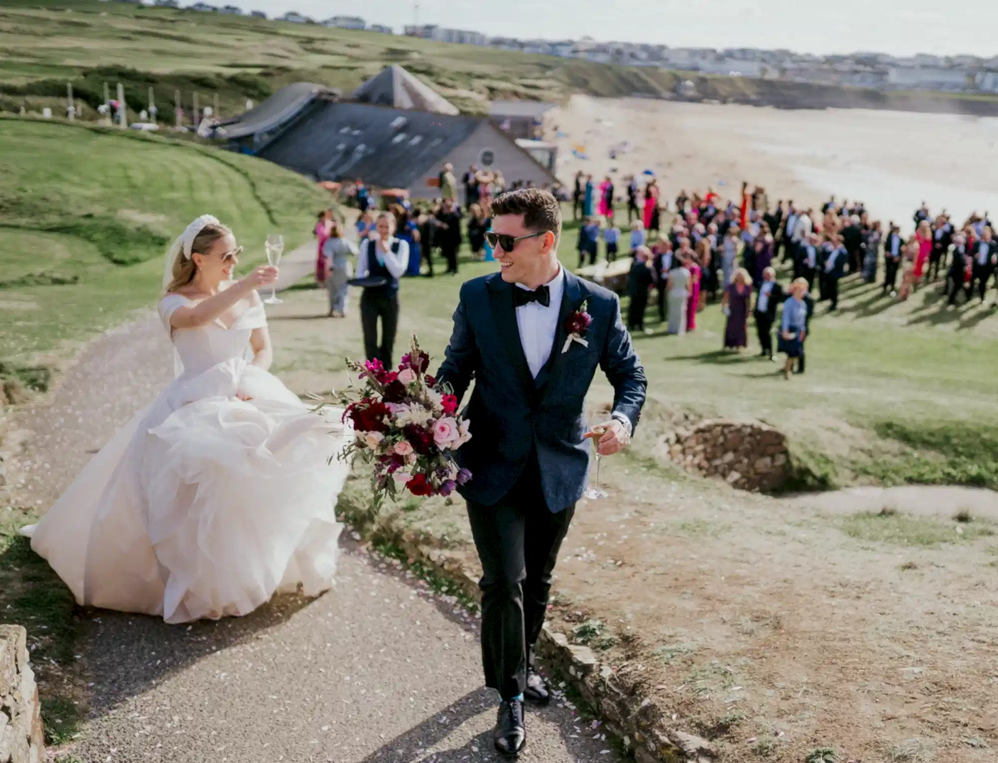 Bride and groom running up a coastal path. The groom is holding a large bouquet of flowers and the bride is toasting with a glass of champagne. The grass lawn is full of guests overlooking Fistral beach and celebrating with the married couple. 