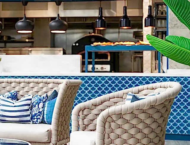 Detail of woven chairs with blue patterned cushions in a luxury Cornwall hotel restaurant, featuring an open-plan kitchen and blue tiled bar in the background.