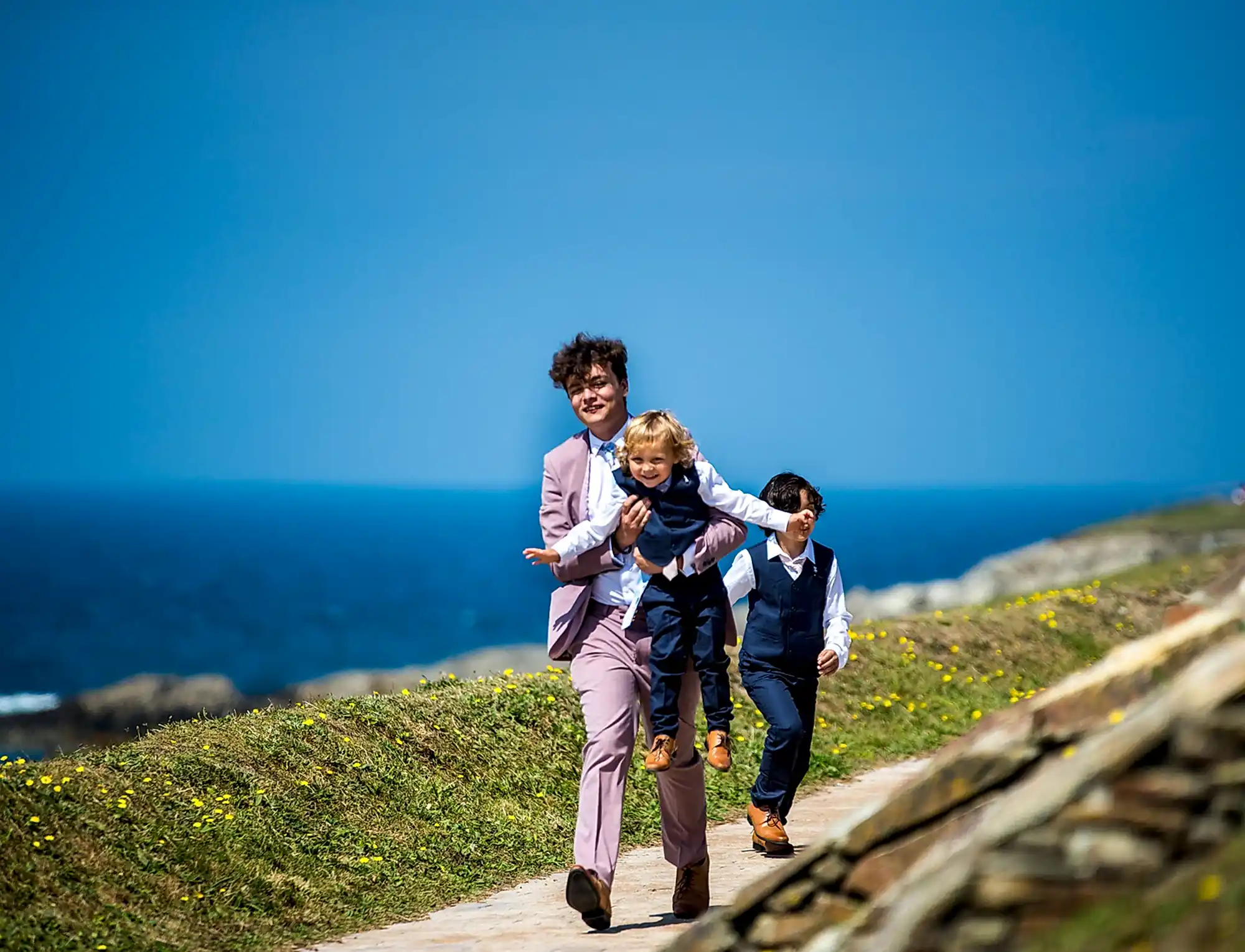 Teen boy along with two younger children walking across a coastal path on a sunny day. All are smartly dressed wearing suits. 