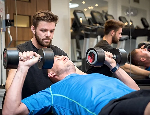 A man doing dumbbell presses whilst being supervised