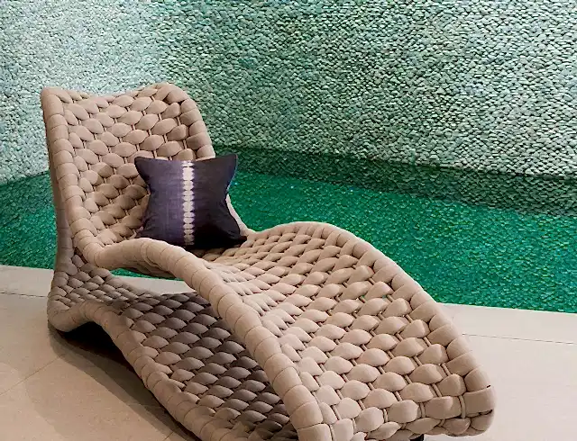 Woven beige lounge chair next to a swimming pool below a curved, stone wall. 